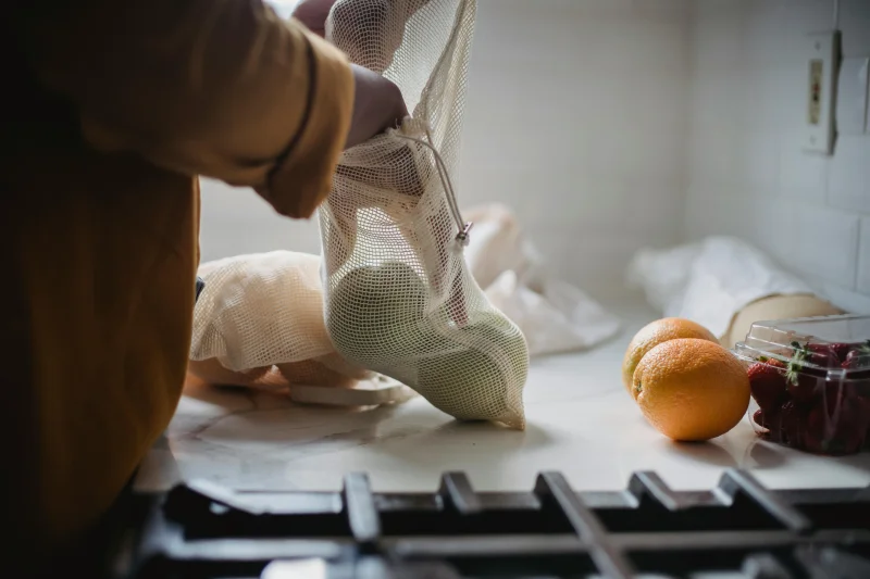Hand placing fresh fruits in a mesh bag on a kitchen counter, promoting sustainability.