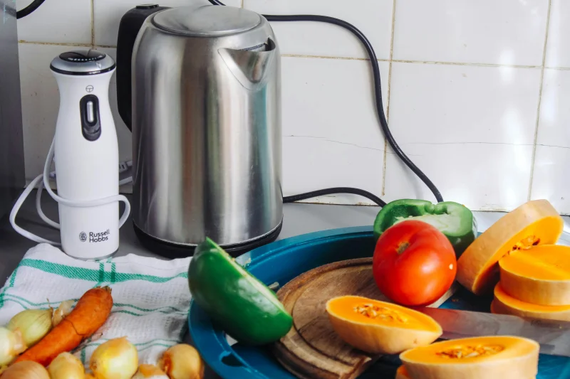 A vibrant array of fresh vegetables with a kettle and a blender on a kitchen countertop, ideal for healthy cooking.