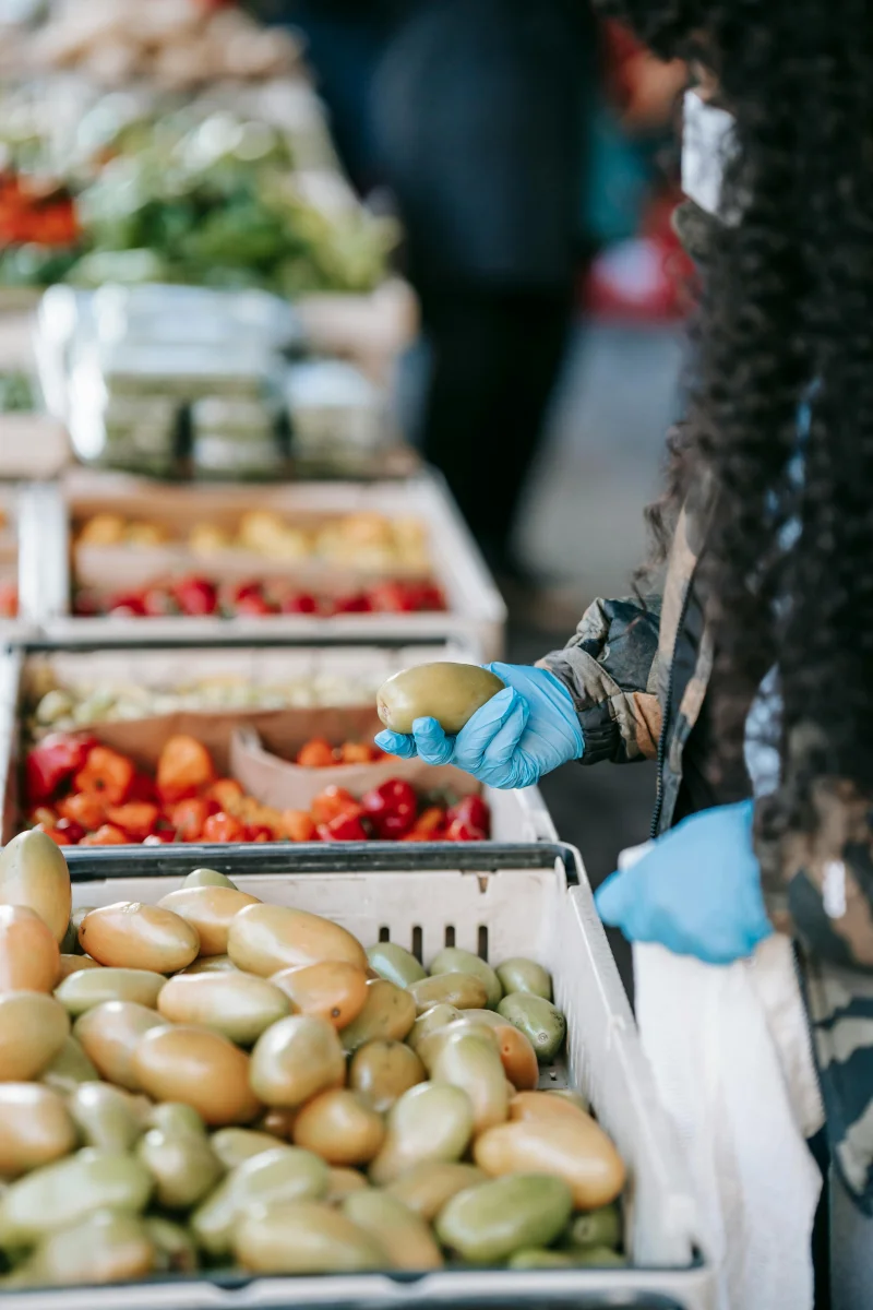 Crop anonymous curly dark haired woman in protective mask and gloves choosing ripe vegetables and putting in plastic bag on street market