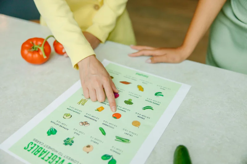 Close-up of hands pointing to a vegetable nutrition chart with fresh tomatoes on the table.