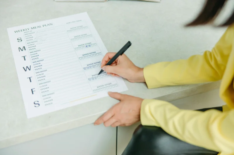 Woman writing on a weekly meal plan checklist indoors, focusing on diet organization.