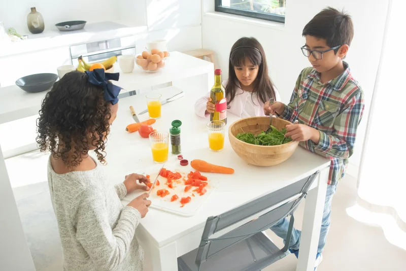 Three children preparing a fresh vegetable salad together in a bright kitchen.