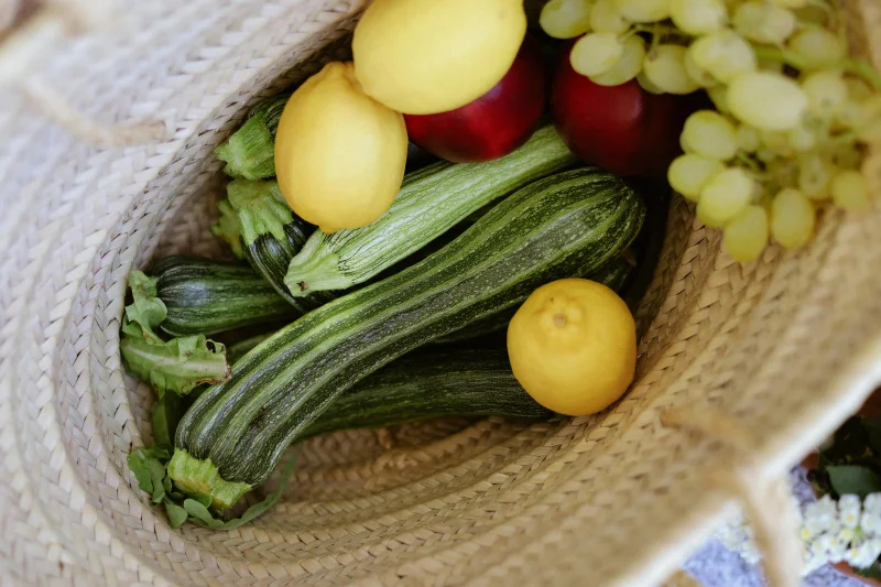 An assortment of zucchini, lemons, red apples, and grapes in a woven basket.