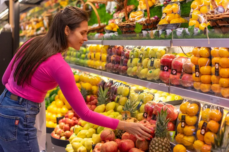 A woman in a bright pink top selecting fresh fruits in a grocery store produce aisle.