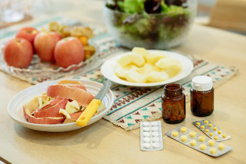 From above of various medicines placed on table near plates with fresh peeled apples and bowl of salad in kitchen