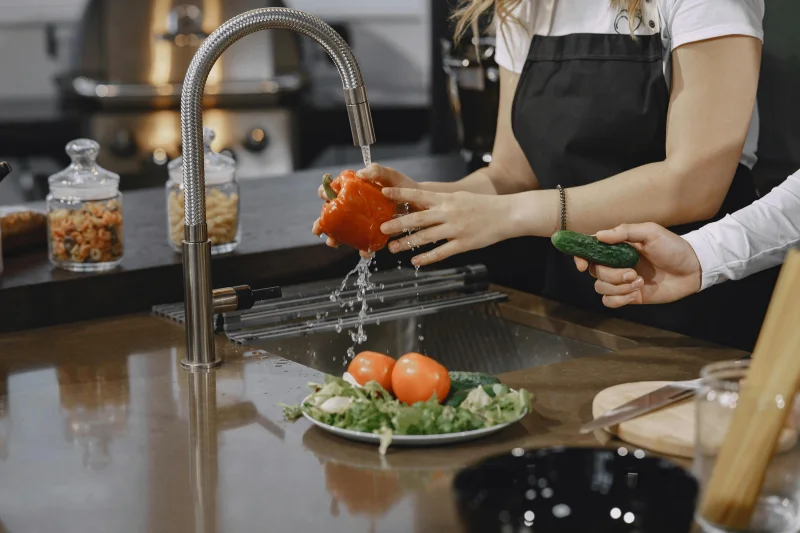 Hands washing a red bell pepper in a modern kitchen sink with fresh vegetables.