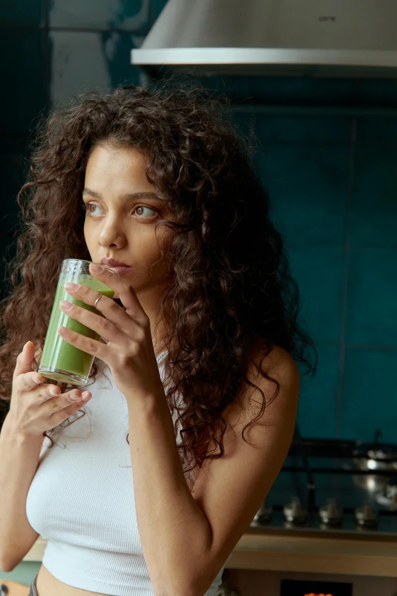 Young woman with curly hair enjoying a green smoothie in a modern kitchen setting.