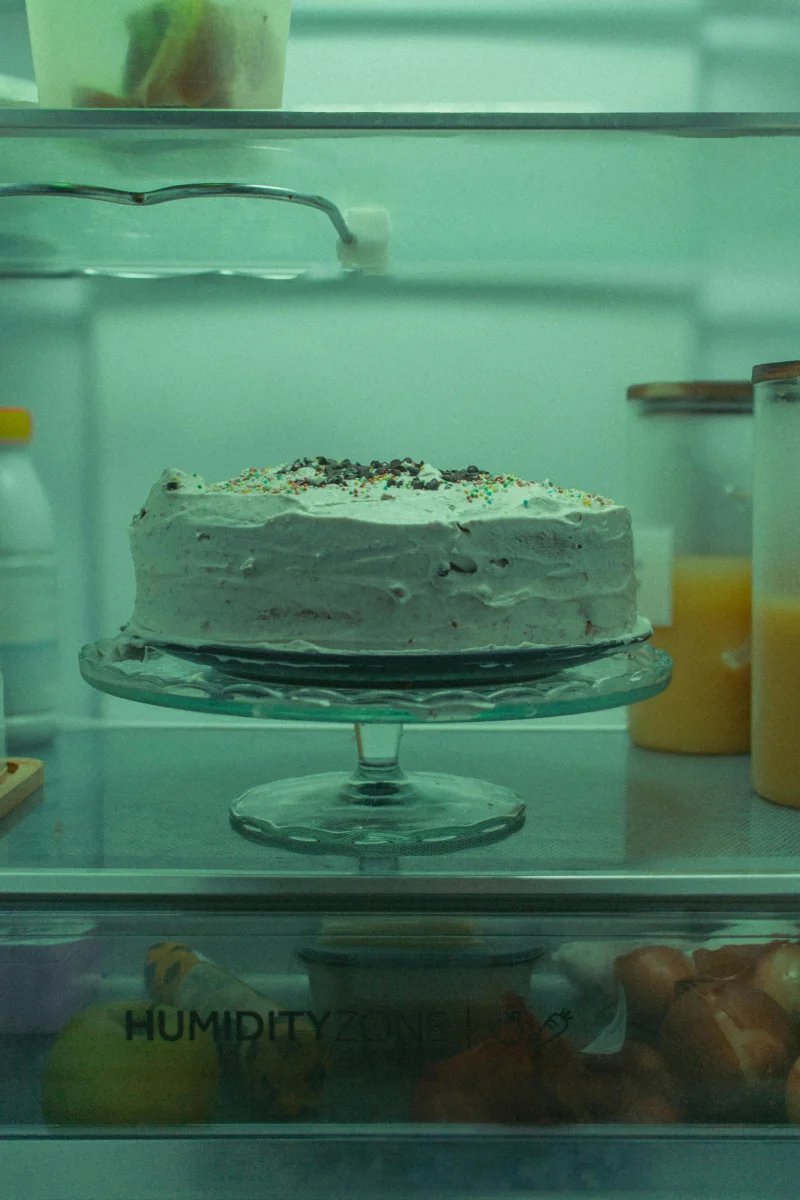 A delicious cake displayed on a stand inside a refrigerator, bathed in a cool green light.