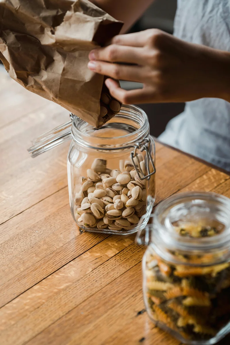 Hands pouring pistachios from brown paper bag into a glass jar on a wooden table.