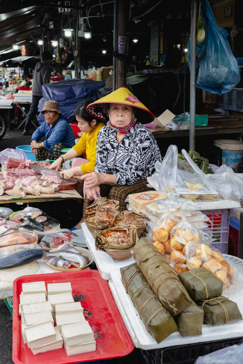 Asian street market vendor with traditional hat selling fresh produce and wrapped foods.