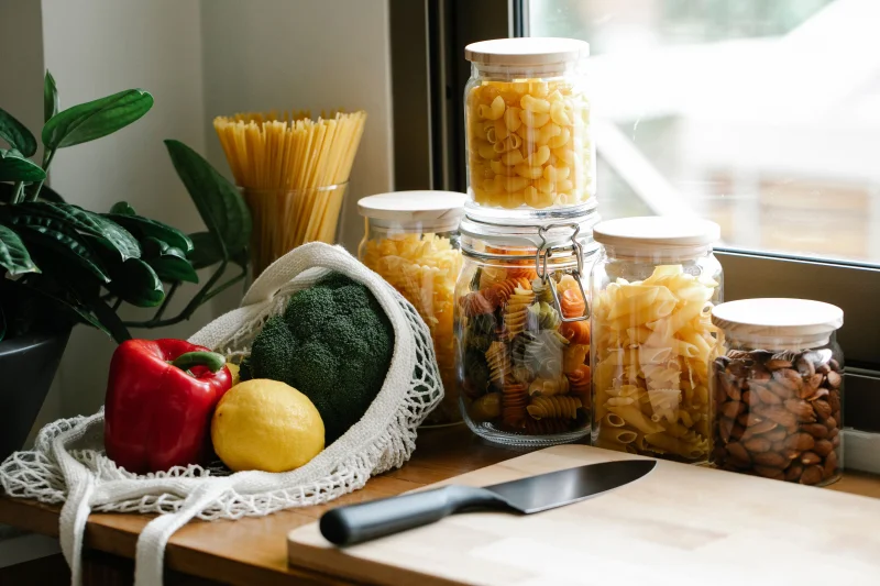 Ripe fresh vegetables in net bag placed on kitchen counter near glass jars with pasta and cutting board with knife