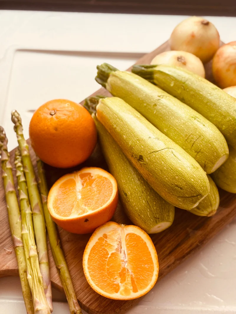 Assorted fresh produce including zucchini, oranges, onions, and asparagus on a wooden board.