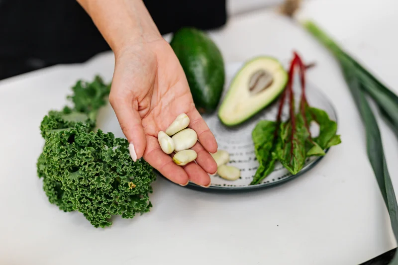 Close-up of a hand holding garlic cloves over a plate with fresh vegetables, including kale and avocado.