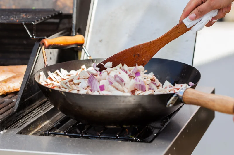 Close-up of hand cooking mushrooms and onions in a pan outdoors with a wooden spatula.