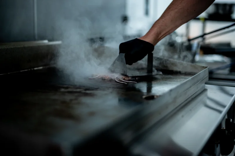 Steam rises as a chef grills meat on a professional griddle in a kitchen.