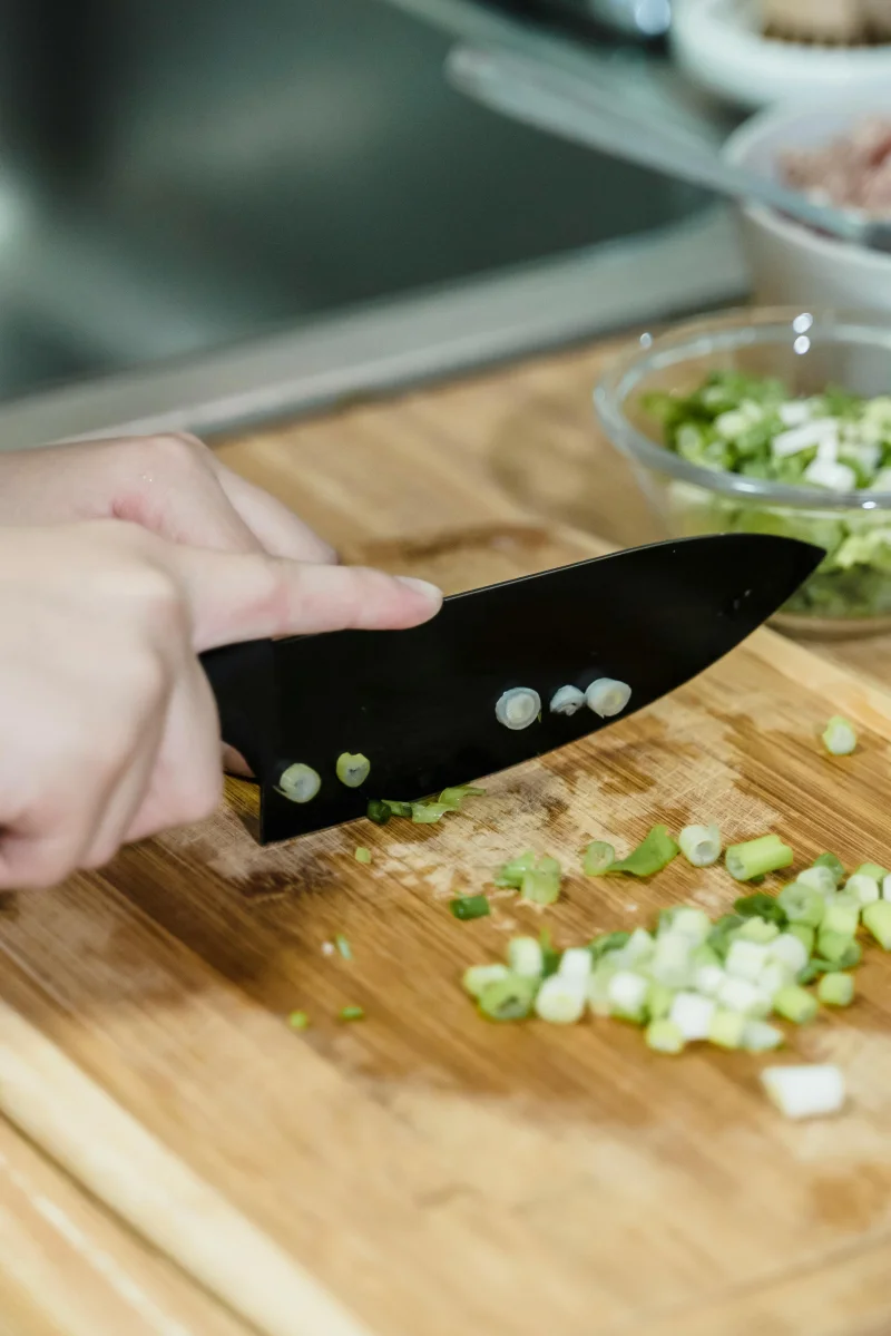 Extreme close-up of hands chopping fresh green onions on a wooden board indoors.