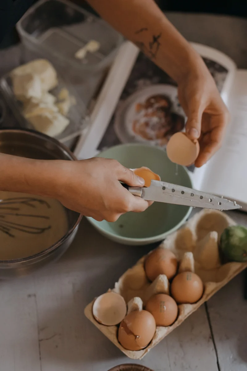A person cracking eggs into a bowl with other ingredients on a kitchen counter.