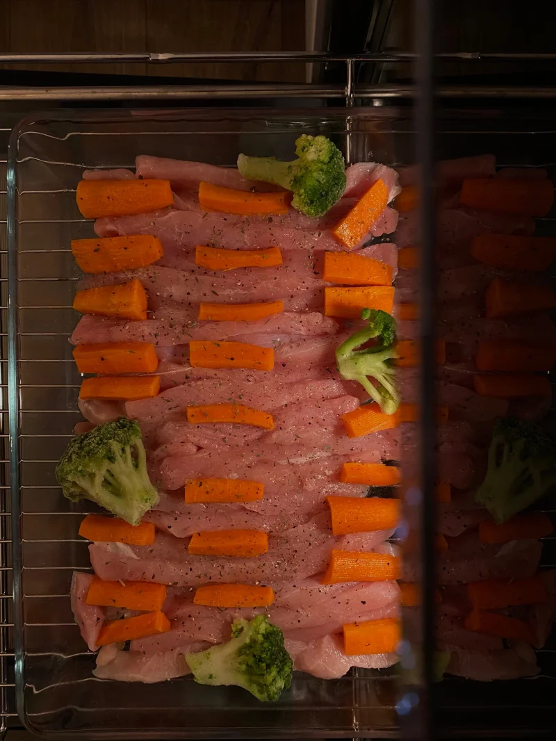 Close-up of raw chicken fillets arranged with carrots and broccoli on a baking tray, ready for cooking.