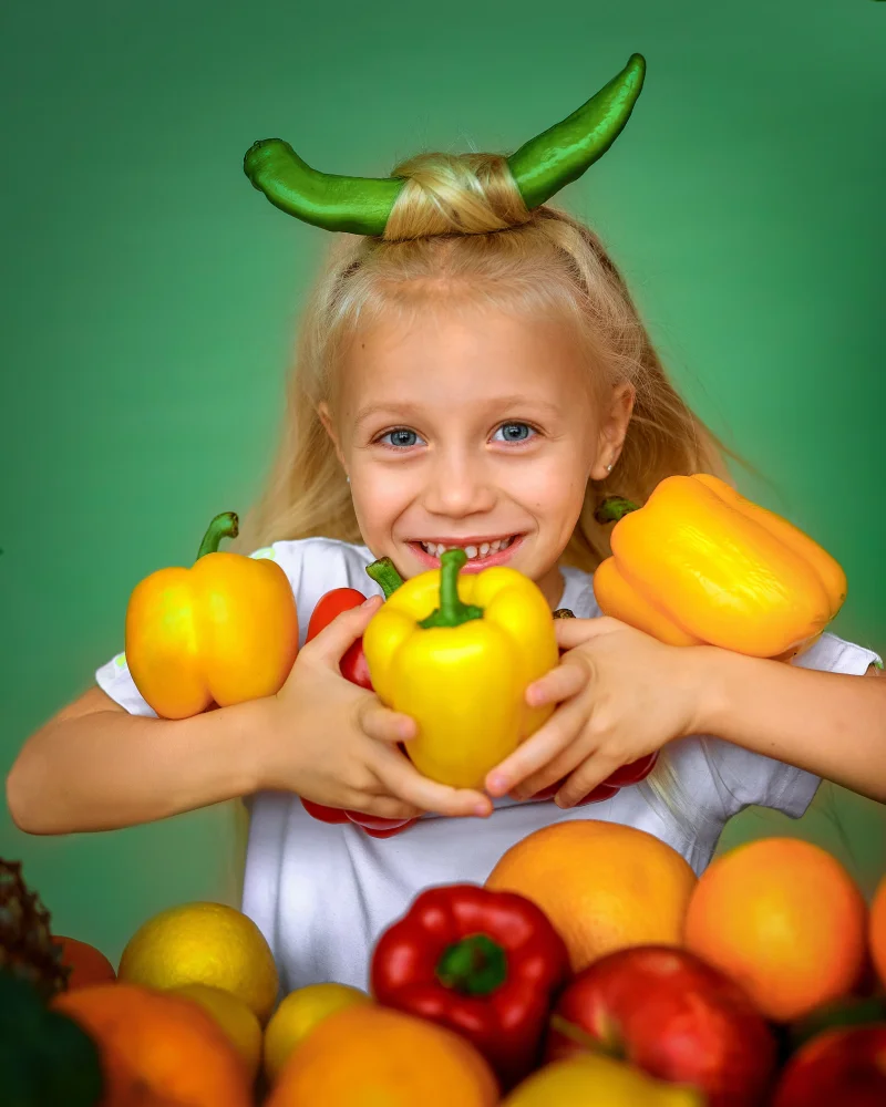 Cheerful blonde girl holding yellow bell peppers with fruits, showing joy and playfulness.