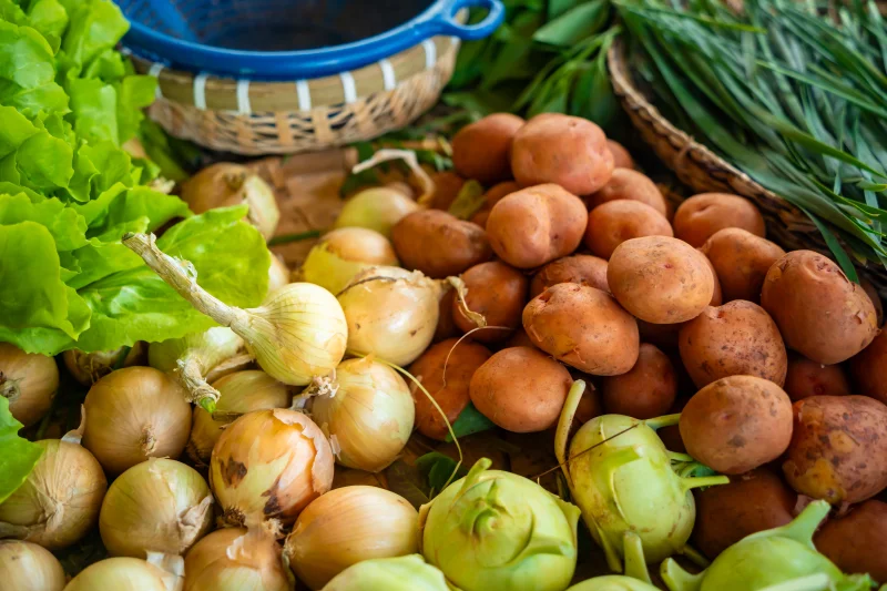 Close-up of assorted fresh vegetables including onions, potatoes, and greens at a farmers market stall.
