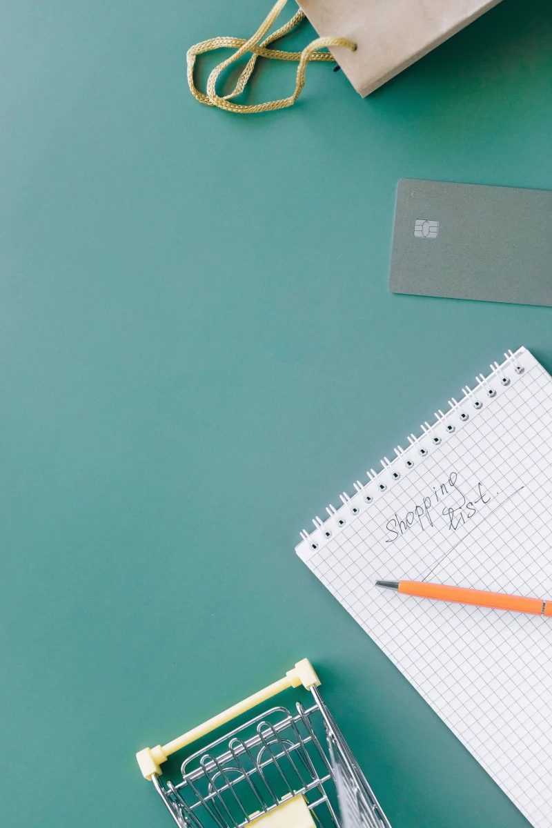 A shopping list on a spiral notebook with a bank card and mini cart on green background.