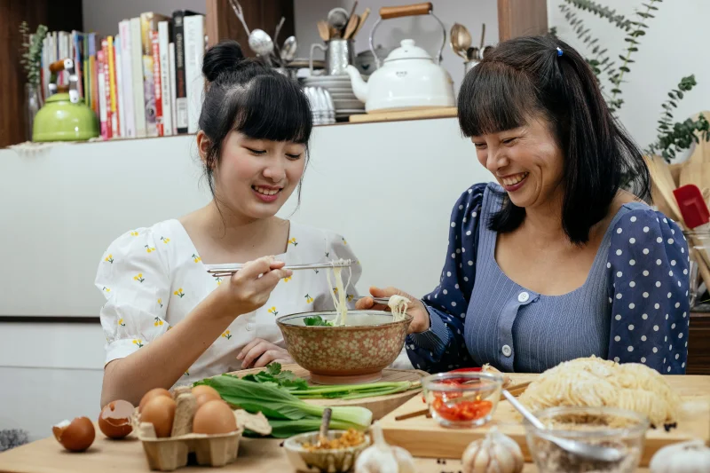 Happy smiling Asian females in casual dresses eating freshly cooked delicious noodles soup with chopsticks while sitting at table with fresh ingredients