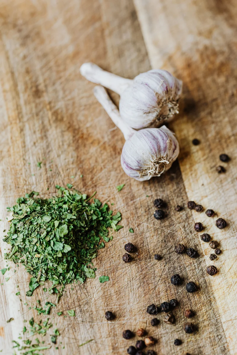 Top-down view of garlic cloves, herbs, and peppercorns on a rustic wooden cutting board.