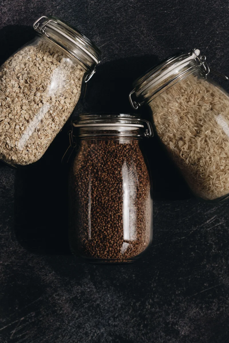 Vertical shot of glass jars with oatmeal, rice, and coffee beans on dark background.