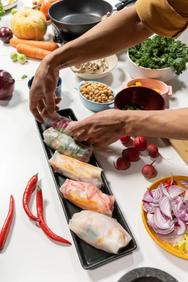 Hands preparing summer rolls with fresh vegetables on a table for a healthy meal.