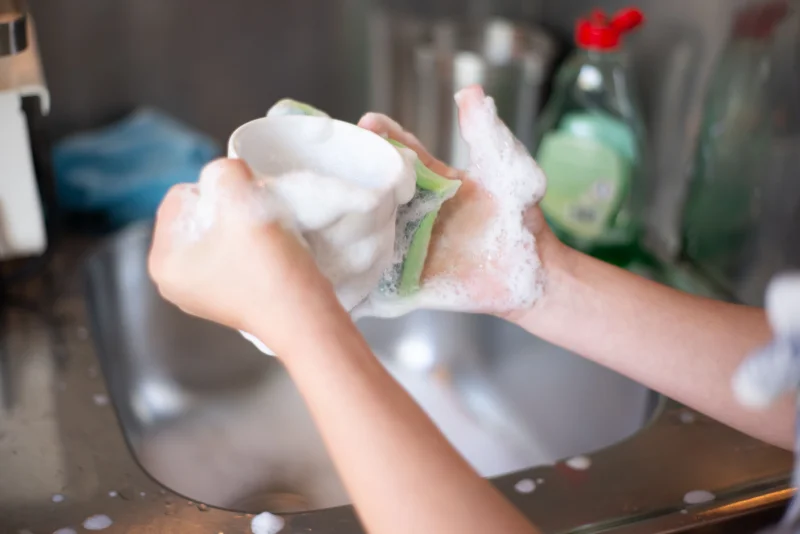 Close-up of hands washing a cup with soap suds in a kitchen sink, representing daily home chores.