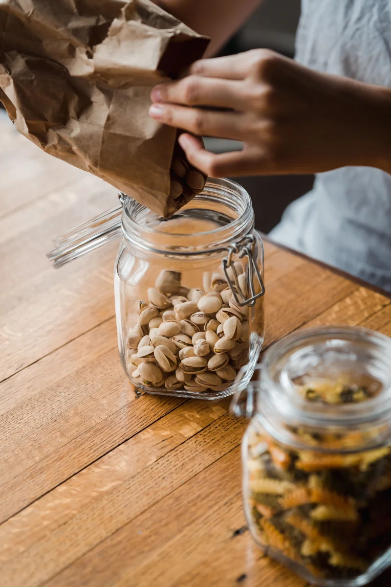 Hands pouring pistachios from brown paper bag into a glass jar on a wooden table.