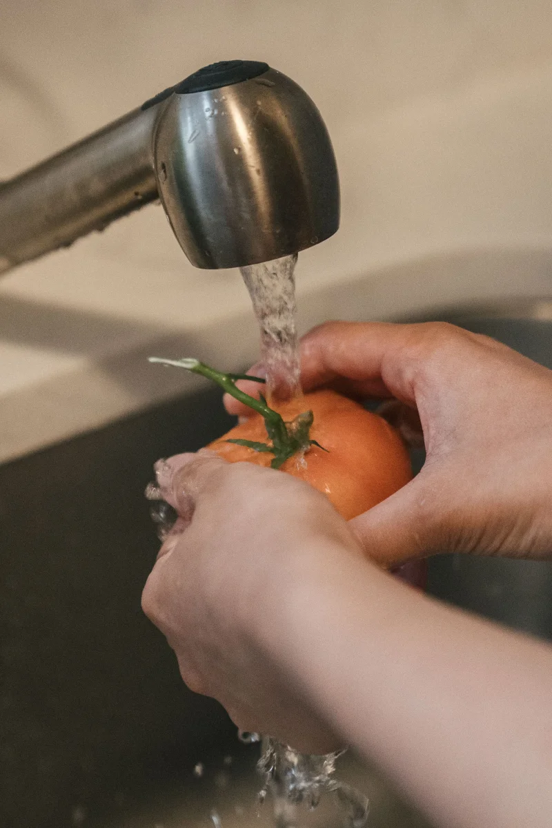 Close-up of hands washing a ripe tomato under a running faucet in a kitchen sink.