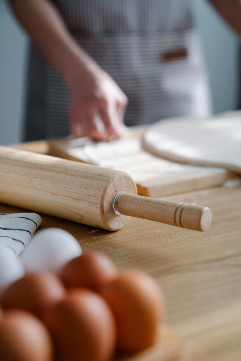 Hands preparing dough with a rolling pin and eggs on a wooden surface in a kitchen setting.