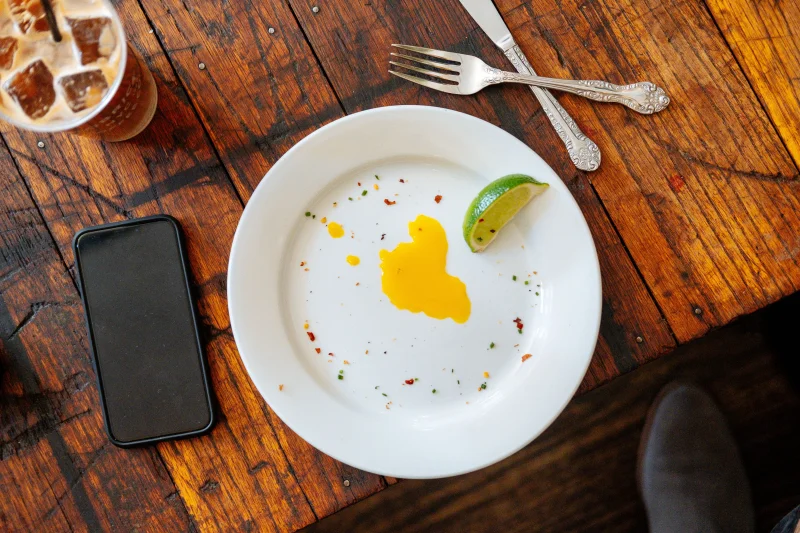 Top view of white plate with slice of lime an egg yolk placed on wooden table near smartphone and iced drink
