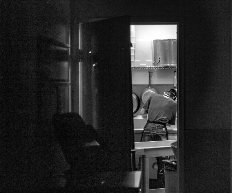 Black and white photo of a dishwasher working in an industrial kitchen setting.