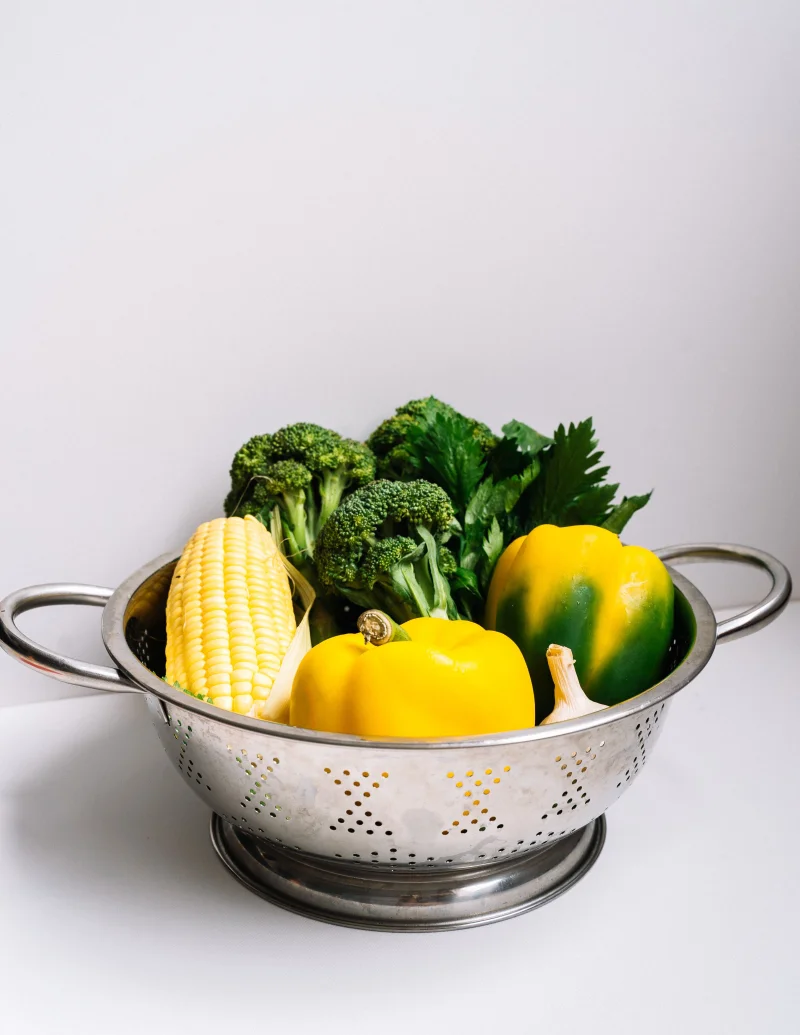 A vibrant collection of fresh vegetables including bell peppers, corn, and broccoli in a stainless steel strainer.