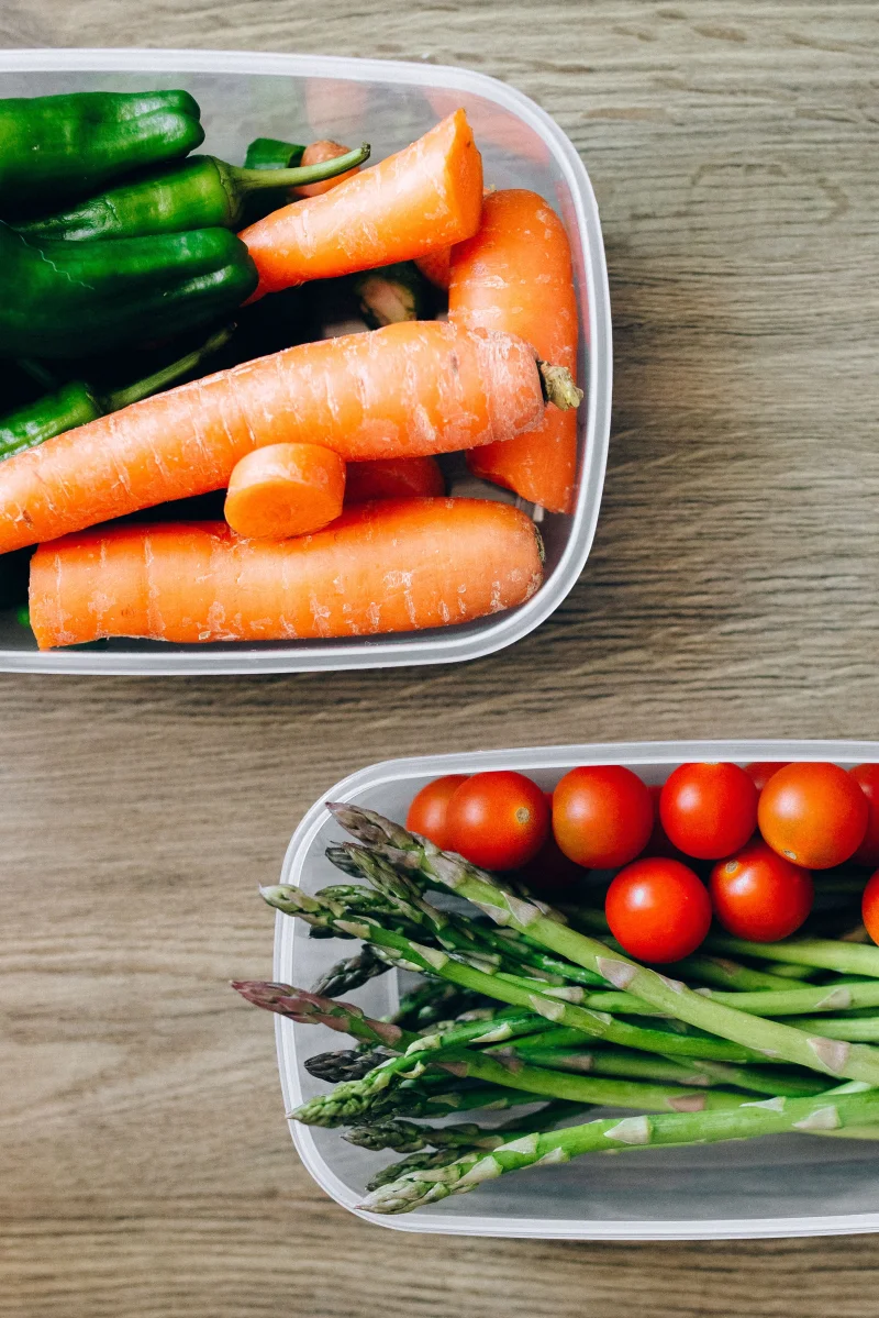 Flat lay of vibrant vegetables including carrots, tomatoes, and asparagus in plastic containers.