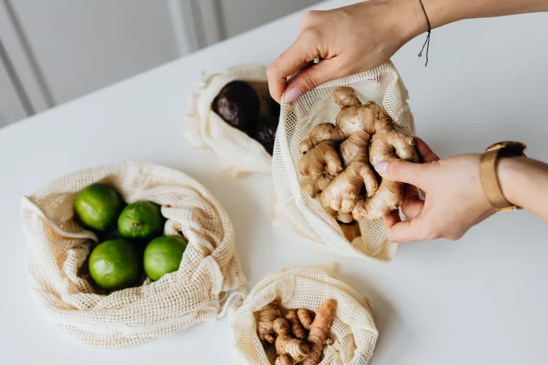 Hands sorting ginger, limes, avocados, and turmeric in reusable net bags on a white surface.