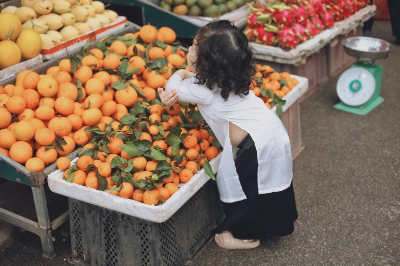 A child in traditional clothing leans over a market stall of fresh oranges.