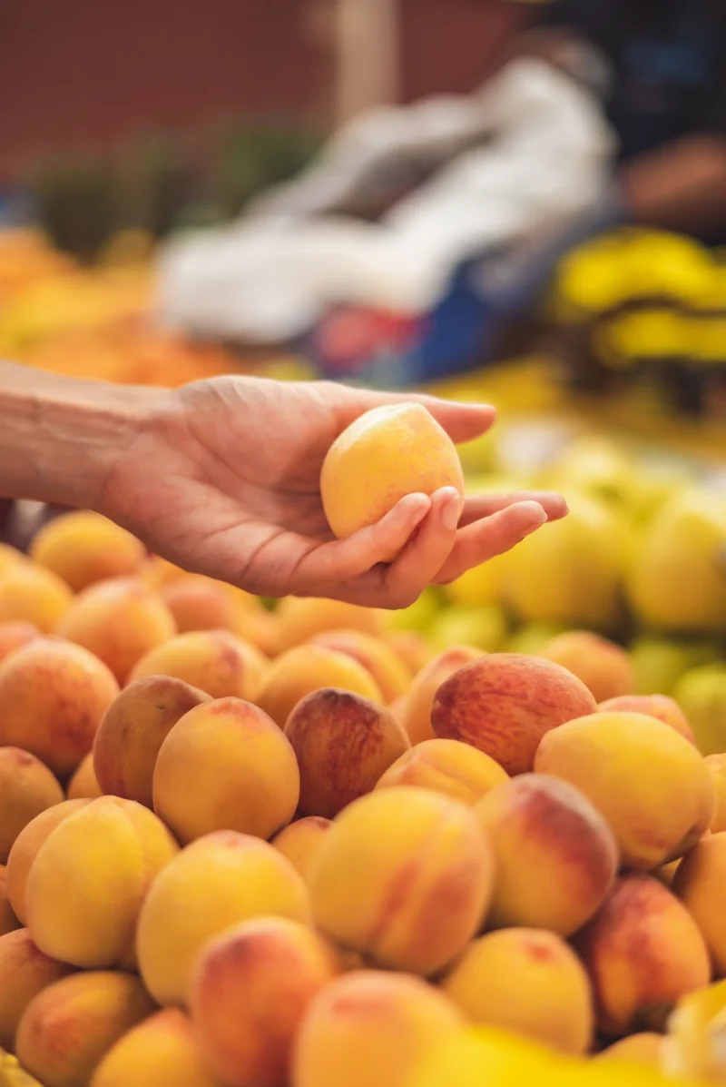 Close-up of a hand selecting a ripe peach in a vibrant market setting.