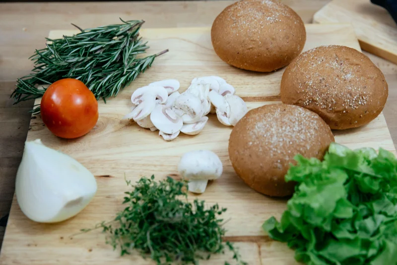 From above of fresh chopped mushrooms with onion and tomato placed on wooden cutting board with assorted  herbs and bread buns during lunch preparation