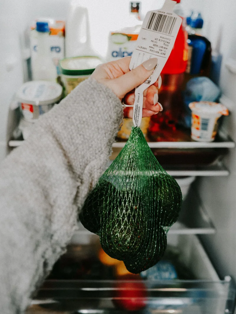 A hand holds a net bag of avocados inside a stocked refrigerator.