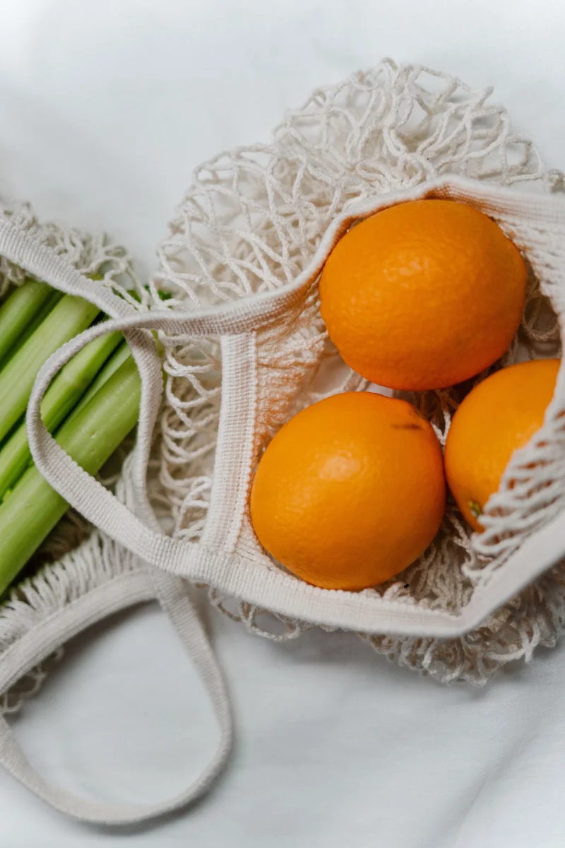 Mesh bags containing fresh oranges and celery stalks, promoting zero waste and eco-friendly living.