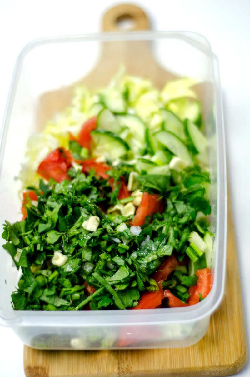 Fresh vegetable salad with cucumbers and herbs in a plastic container on a wooden board.