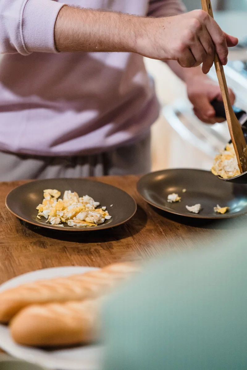 A man serves freshly cooked scrambled eggs onto plates in a cozy kitchen setting.