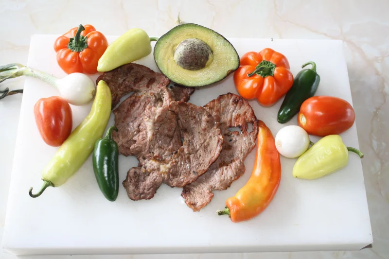A colorful assortment of grilled steak and fresh vegetables on a white cutting board.