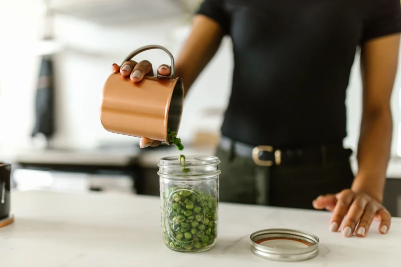 A woman pours green peas into a glass jar on a kitchen counter, emphasizing organization.