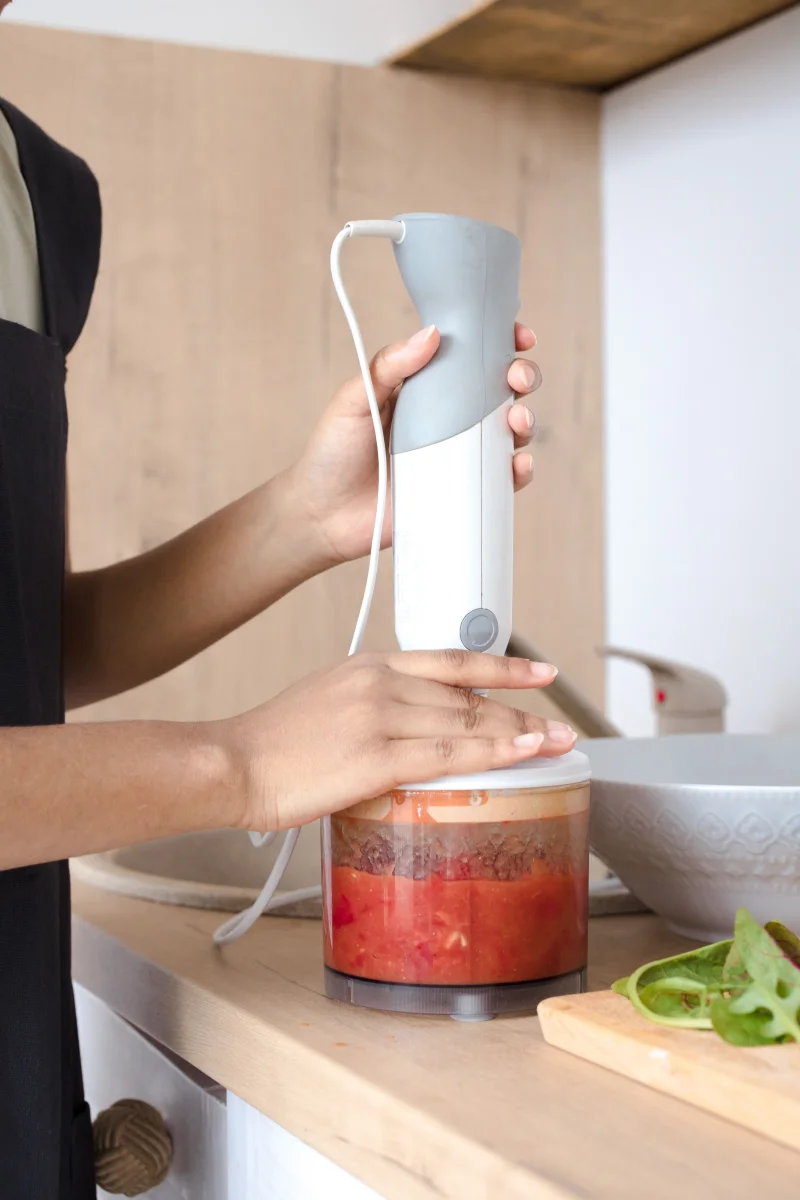 A person blending vegetables with a hand blender in a modern kitchen setting.